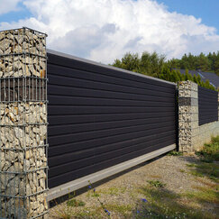 Velký šedý drcený vápencový gabionový plot s vrstvou tmavé skály po obvodu plotu.